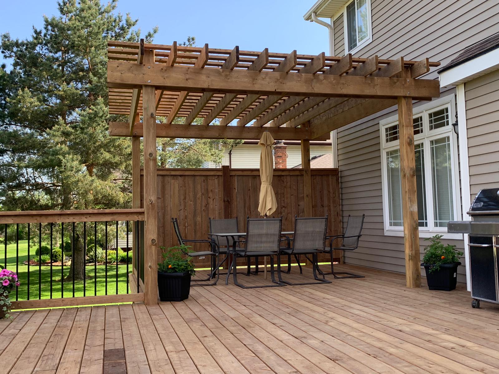 A wooden deck with a pergola, outdoor furniture, and a grill, surrounded by greenery and attached to a house with large windows.