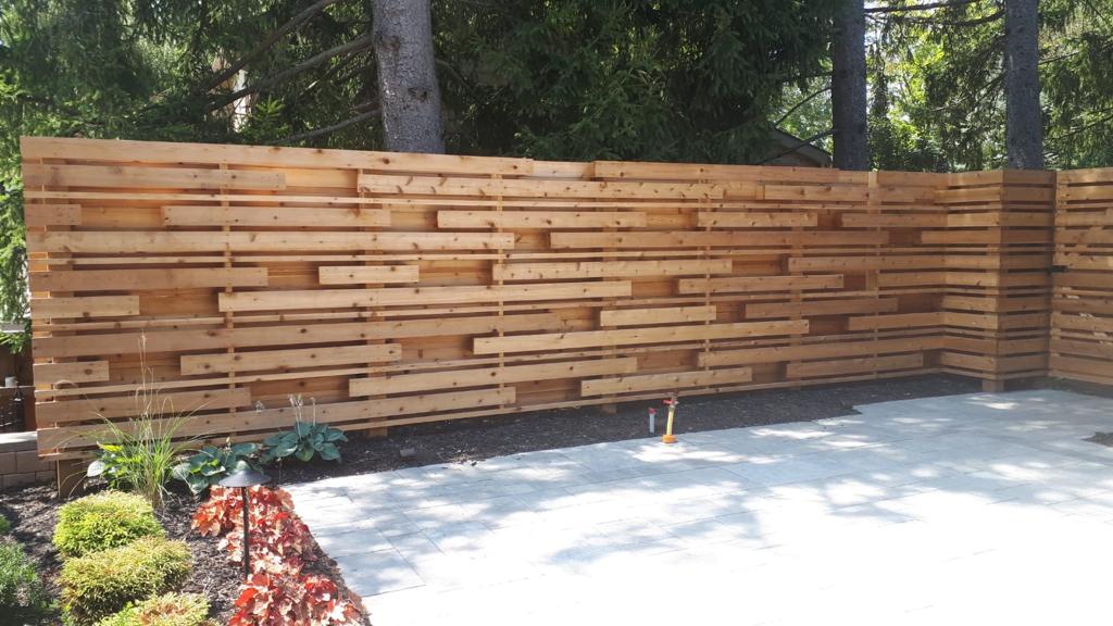 A wooden privacy fence in a garden with landscaped plants, trees, and a paved patio area under natural sunlight.