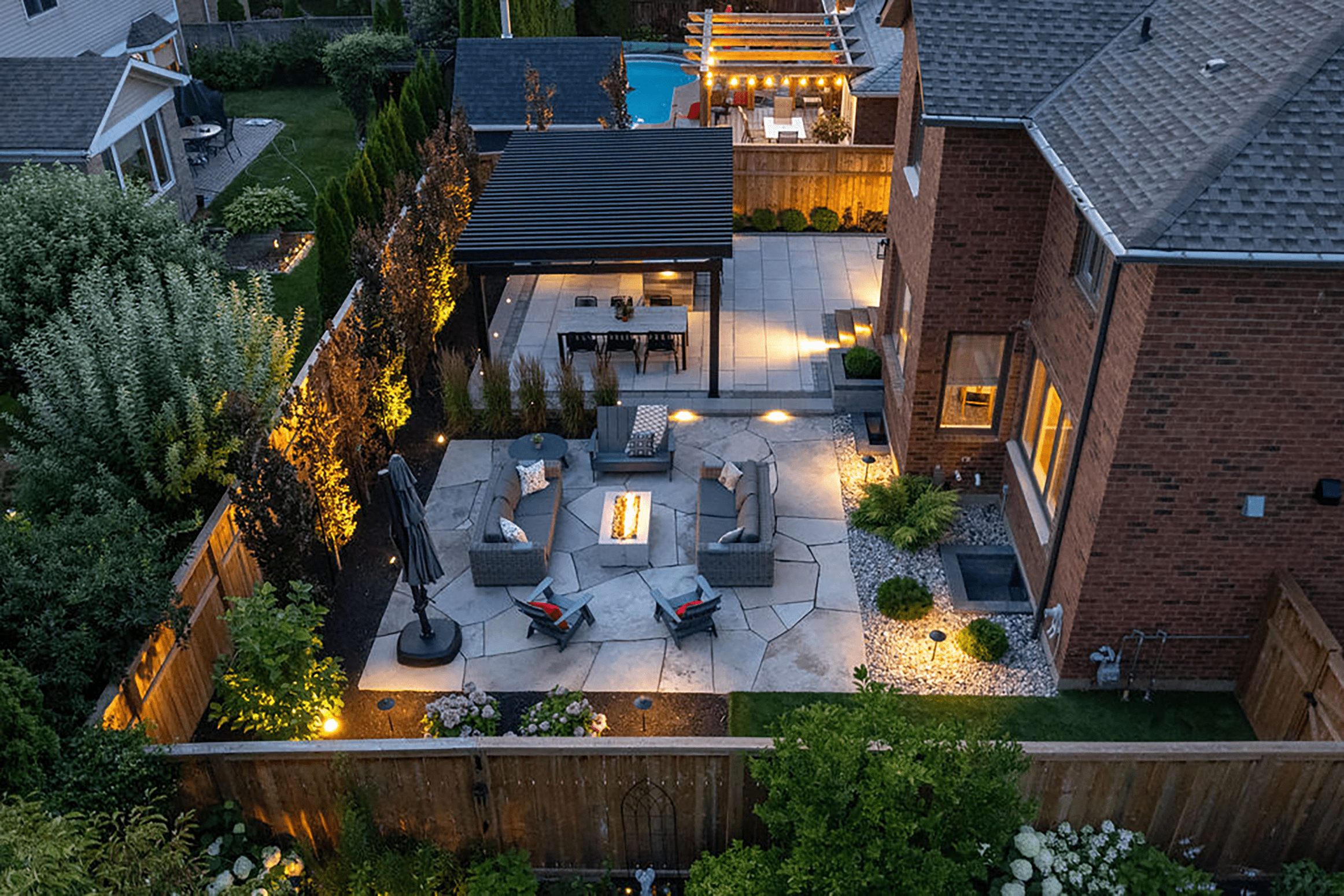Aerial view of a beautifully lit backyard with patio, outdoor seating, fire pit, and landscaped garden adjoining a brick house.