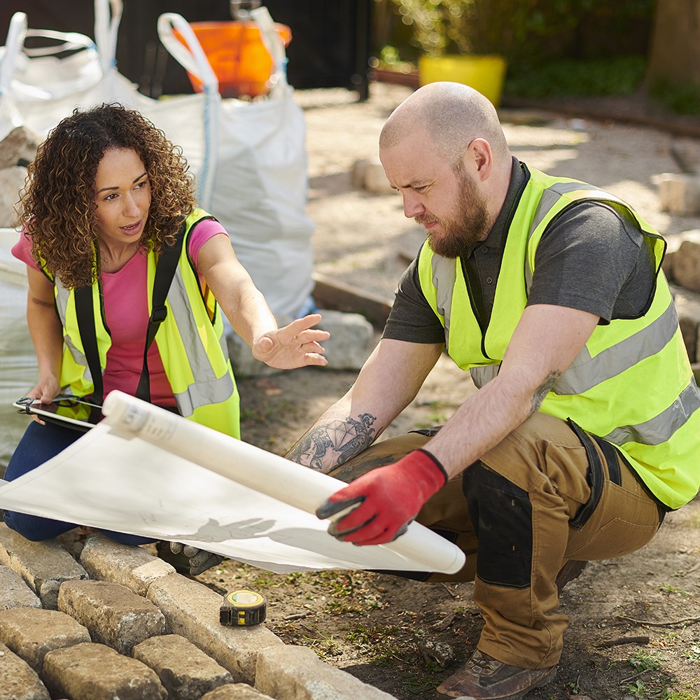 Two persons wearing reflective vests discuss construction plans at a site, surrounded by building materials and tools on a sunny day.